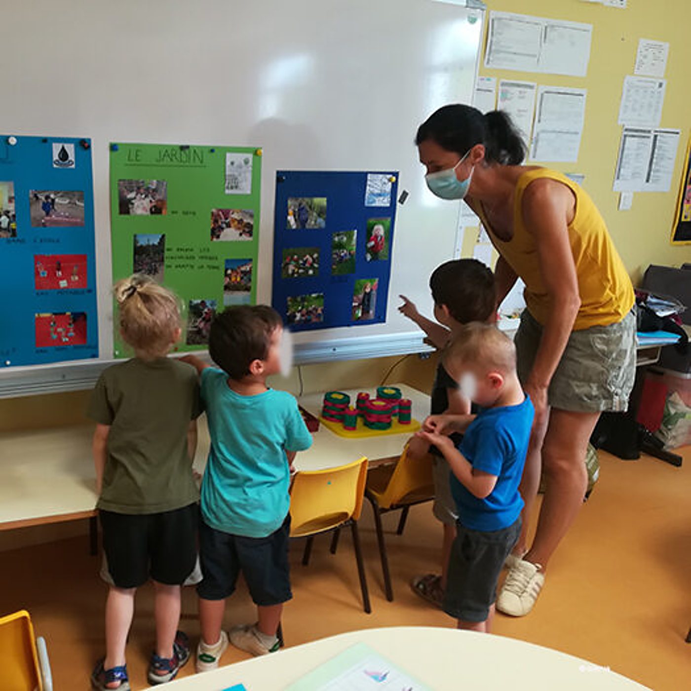 Dans une salle de classe, une éducatrice portant un masque observe quatre jeunes enfants debout devant des panneaux d'affichage dont l'un est intitulé « Le jardin ».