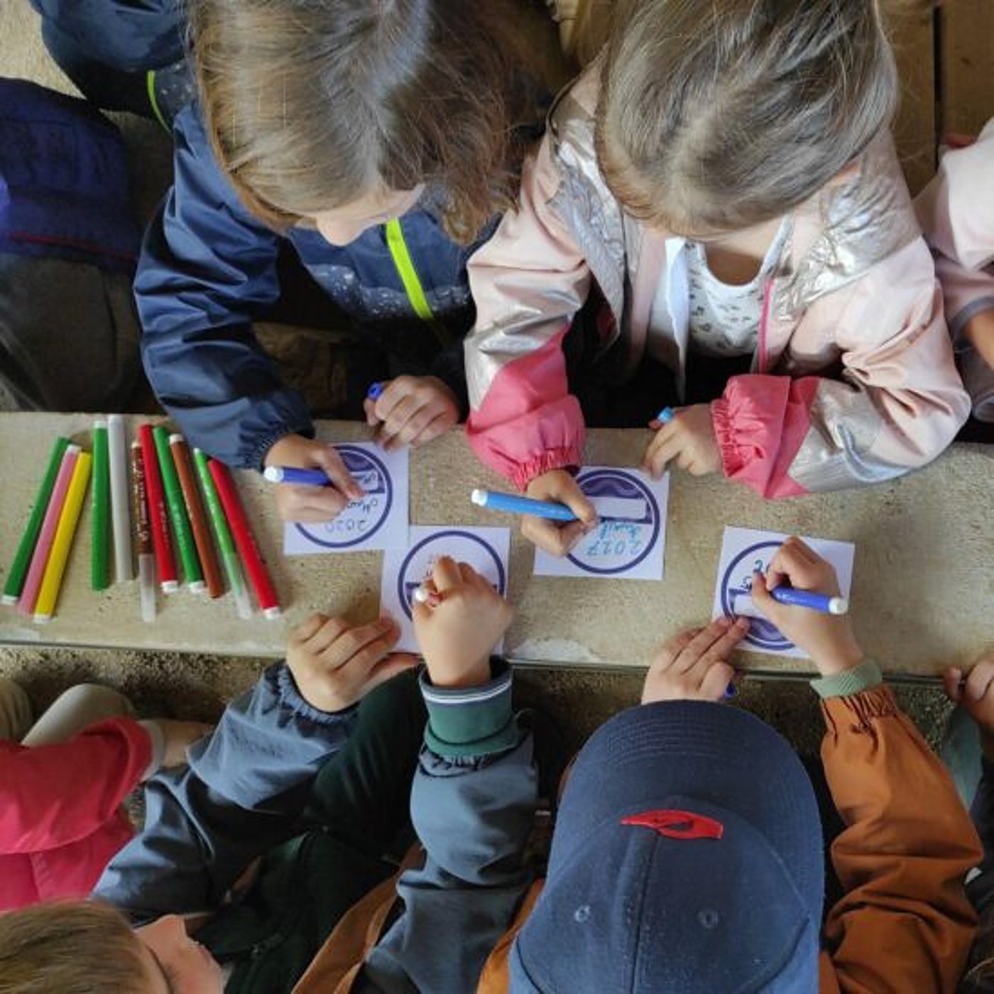 Vue du dessus d'enfants en vêtements d'extérieur qui colorient de petits badges ronds avec des feutres colorés sur une table en pierre.