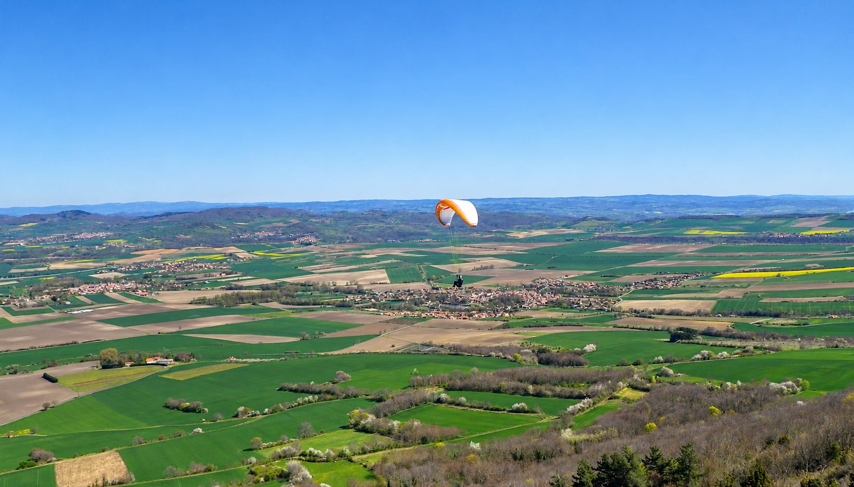 Un parapente orange et blanc vole au-dessus d'une large plaine de champs verts et de villages, sous un ciel bleu dégagé.