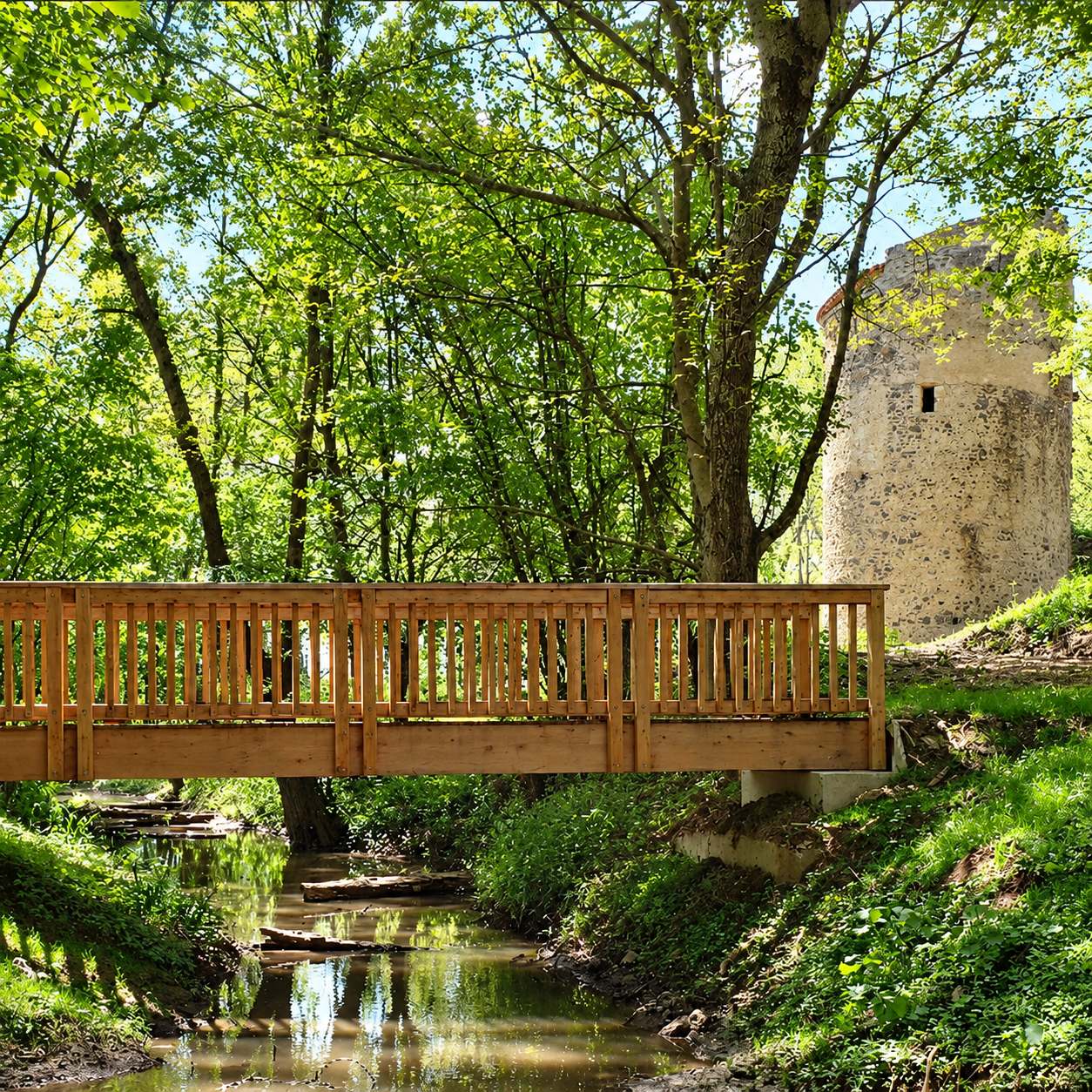 Une passerelle en bois clair avec garde-corps enjambe un ruisseau bordé d'arbres verdoyants, près d'une ancienne tour de pierre.