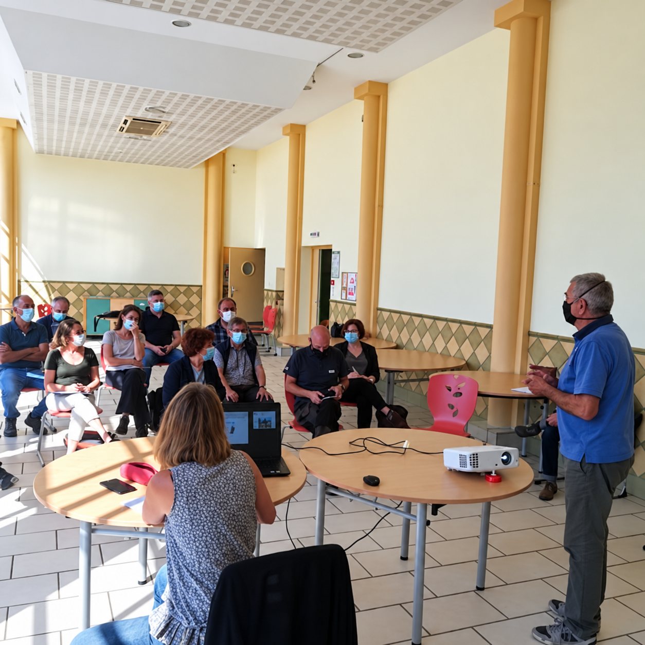 Un homme masqué debout présente un projet à un groupe de personnes masquées assises dans une grande salle lumineuse, avec un vidéoprojecteur posé sur une table ronde au premier plan.